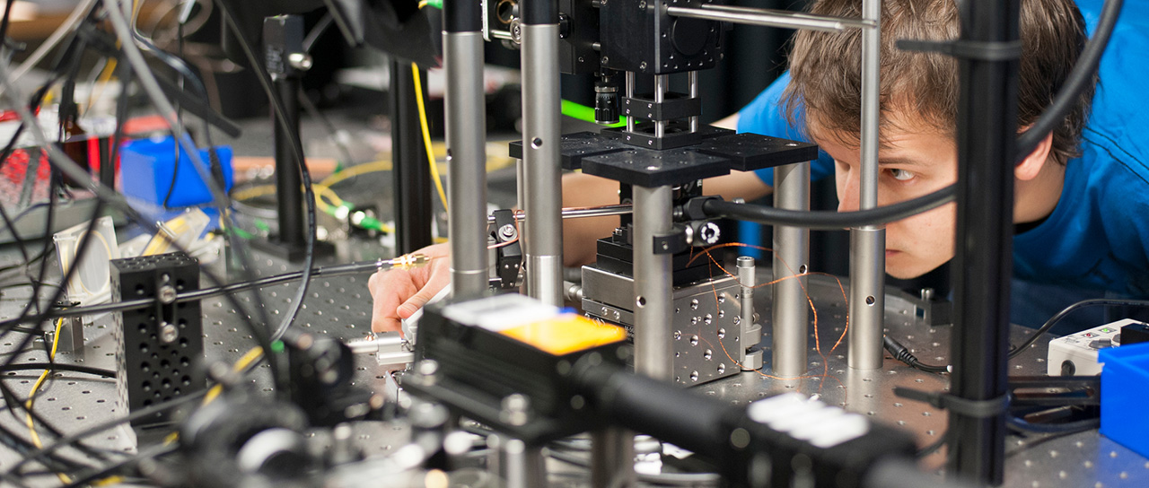 A researcher in a blue shirt is adjusting an optical setup in a laboratory. The setup consists of various optical components, metal frames, and numerous cables. The researcher is carefully aligning the equipment while closely observing the setup.