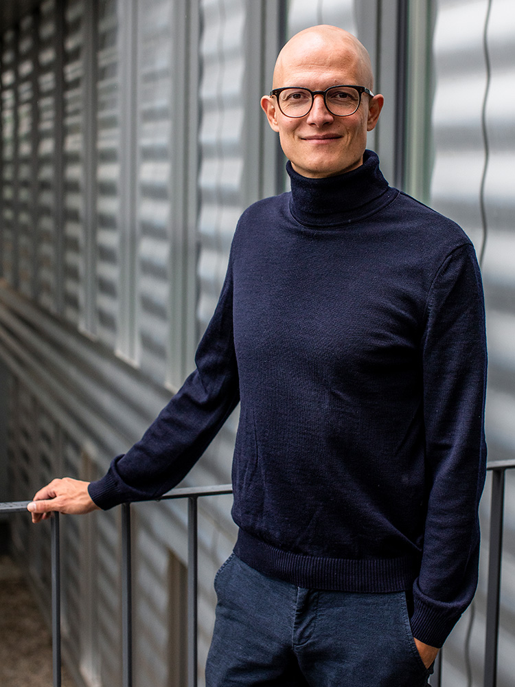 Man with glasses and a shaved head wearing a dark blue turtleneck sweater and dark trousers, standing outdoors in front of a modern, corrugated metal wall. Man with glasses and a shaved head wearing a dark blue turtleneck sweater and dark trousers, standing outdoors in front of a modern, corrugated metal wall.