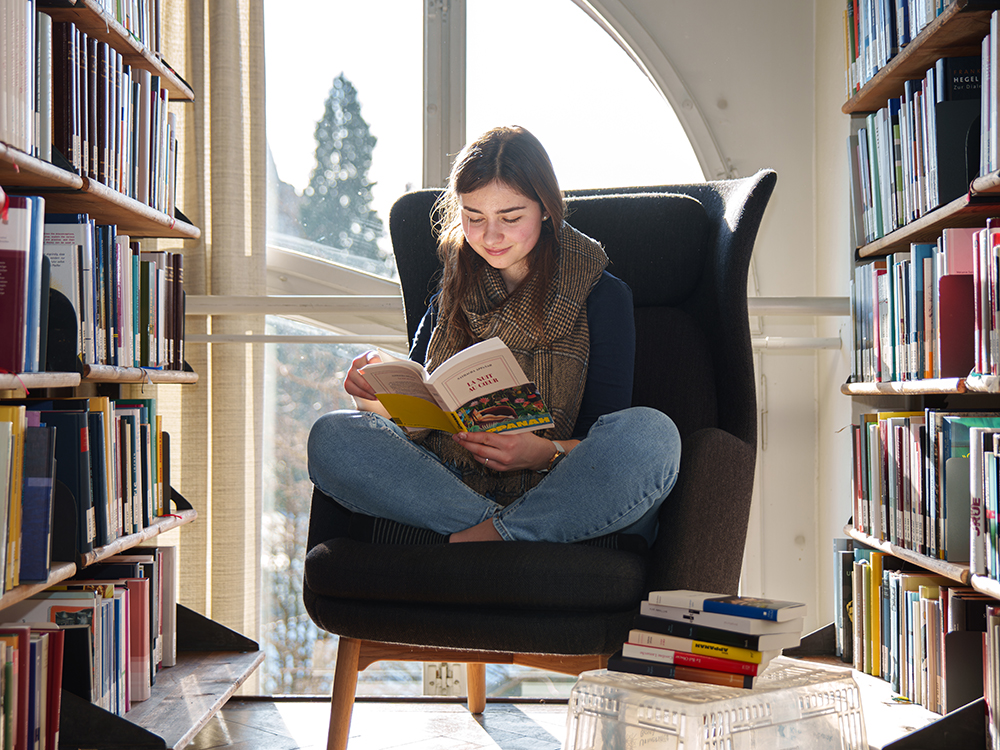 Junge Frau sitzt in einem Sessel in einer Bibliothek und liest ein Buch, umgeben von Bücherregalen und beleuchtet durch Tageslicht aus einem grossen Fenster. Junge Frau sitzt in einem Sessel in einer Bibliothek und liest ein Buch, umgeben von Bücherregalen und beleuchtet durch Tageslicht aus einem grossen Fenster.