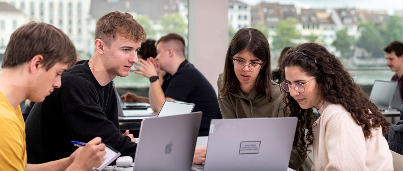 Eine Gruppe von Studierenden sitzt an einem Tisch und arbeitet an Laptops. Zwei Frauen und ein Mann arbeiten zusammen, während ein weiterer Mann Notizen macht. Im Hintergrund sind weitere Studierende bei der Arbeit zu sehen. Der Raum hat grosse Fenster mit Blick auf Gebäude und den Rhein.