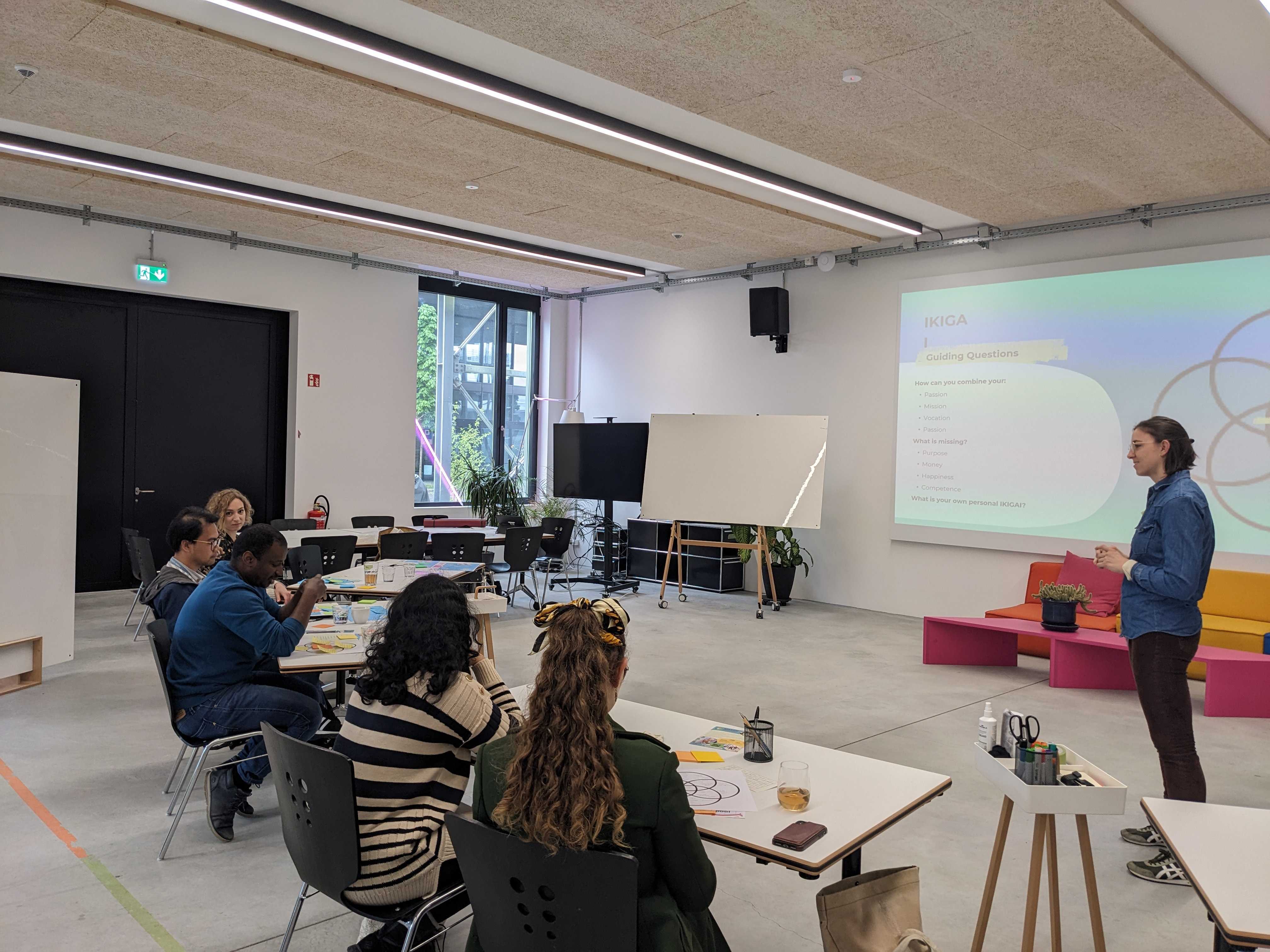 Ein Seminarraum, mehrere Tische im Halbkreis, ausgerichtet auf die Person in der Mitte und die Powerpoint-Präsentation hinter hier. Ein Seminarraum, mehrere Tische im Halbkreis, ausgerichtet auf die Person in der Mitte und die Powerpoint-Präsentation hinter hier.