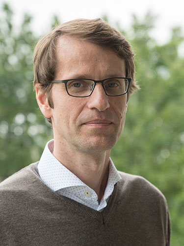 Porträt eines Mannes mit kurzen braunen Haaren und Brille, der ein weißes Hemd mit dezentem Muster und einen braunen Pullover trägt, vor einem unscharfen grünen Hintergrund im Freien. Porträt eines Mannes mit kurzen braunen Haaren und Brille, der ein weißes Hemd mit dezentem Muster und einen braunen Pullover trägt, vor einem unscharfen grünen Hintergrund im Freien.