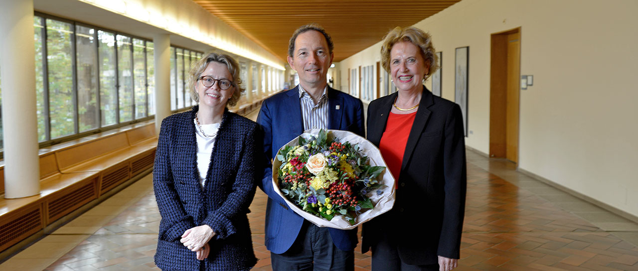 A man holding a bouquet of flowers stands between two women in a hallway with large windows. All three are smiling and dressed in business attire. The hallway has a wooden ceiling and a tiled floor.