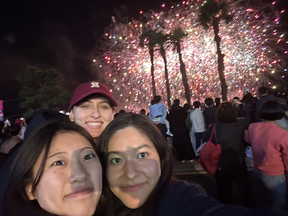 Anna und ihre beiden Freundinnen machen ein Selfie vor einer Menschenmenge, die bunte Feuerwerke in der Nacht beobachtet. Anna und ihre beiden Freundinnen machen ein Selfie vor einer Menschenmenge, die bunte Feuerwerke in der Nacht beobachtet.