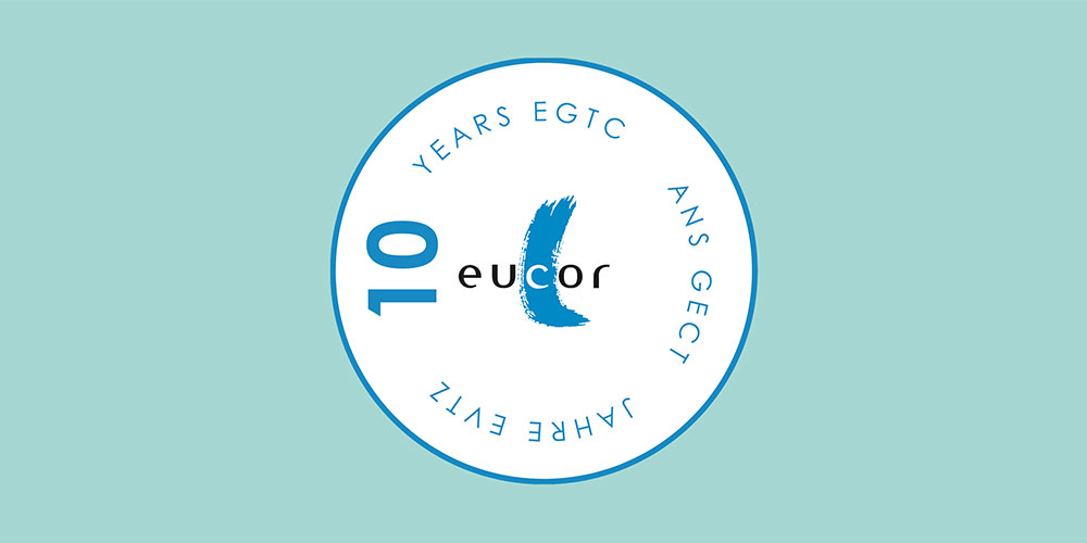 Circular logo on a light blue background with the text "10 Years EGTC / 10 Ans GECT / 10 Jahre EVTZ" around the edge and "eucor" in the center with a blue brushstroke design. Circular logo on a light blue background with the text "10 Years EGTC / 10 Ans GECT / 10 Jahre EVTZ" around the edge and "eucor" in the center with a blue brushstroke design.