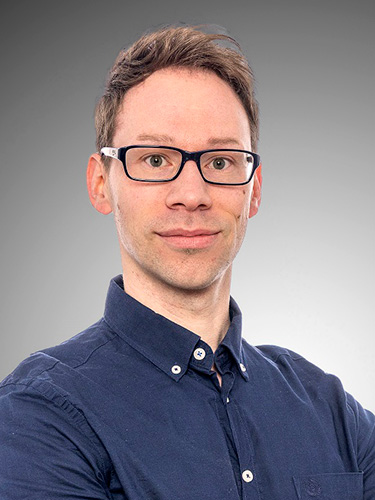 A portrait of a man with short brown hair, black glasses, and a dark blue shirt. A portrait of a man with short brown hair, black glasses, and a dark blue shirt.