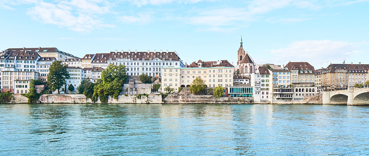 Uferansicht von historischen Gebäuden in Basel mit der Alten Universität am Rhein unter blauem Himmel.