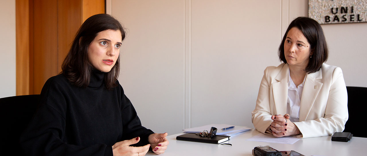 Two women seated at a table in an office setting, engaged in a conversation. One woman wears a black sweater, gesturing with her hands, while the other wears a white blazer, listening attentively.