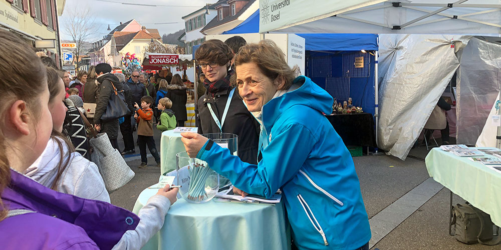 A woman in a blue jacket speaks to a young girl at an outdoor event. They stand near a table with brochures under a tent from the University of Basel. A crowd of people is visible in the background, along with market stalls. A woman in a blue jacket speaks to a young girl at an outdoor event. They stand near a table with brochures under a tent from the University of Basel. A crowd of people is visible in the background, along with market stalls.