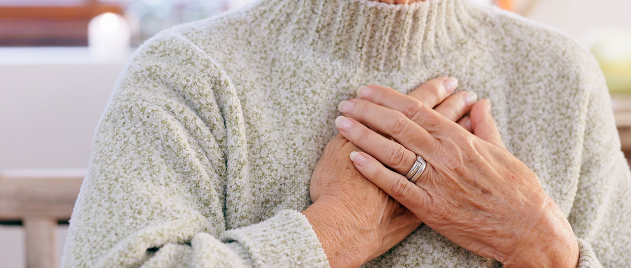 Eine ältere Frau trägt einen grob gestrickten Pullover und hält sich mit beiden Händen die Brust. Die rechte Hand trägt einen silbernen Ring.