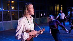 Eine junge Frau in weißer Bluse spielt Ukulele und singt, während im Hintergrund Tänzerinnen und Tänzer auf einer schwach beleuchteten Bühne auftreten.