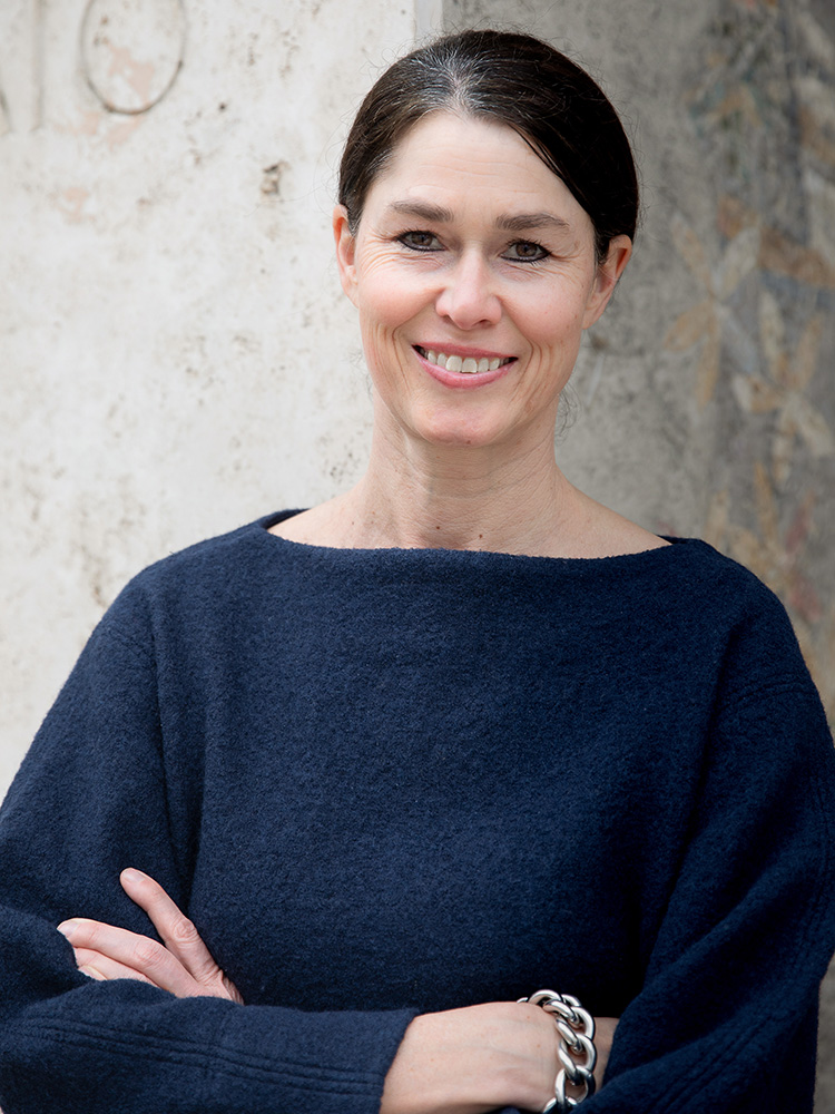 A woman with dark, tied-back hair wears a dark blue sweater and smiles. She is standing in front of a large, slightly blurred mosaic wall with floral and natural motifs. A woman with dark, tied-back hair wears a dark blue sweater and smiles. She is standing in front of a large, slightly blurred mosaic wall with floral and natural motifs.