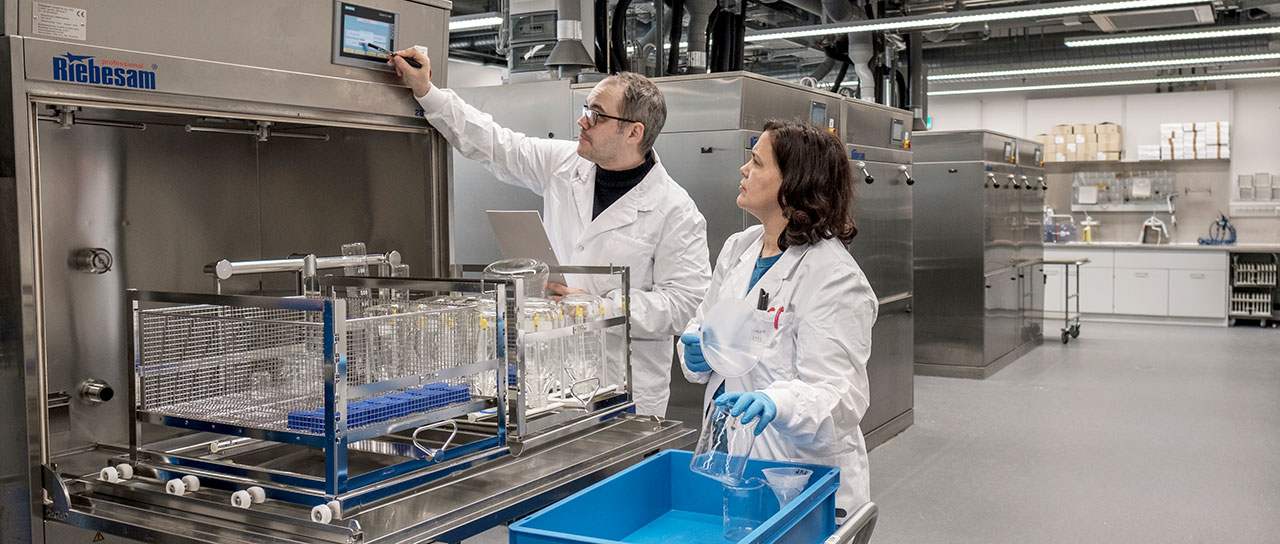 Zwei Personen in weißen Laborkitteln arbeiten mit industriellen Spülmaschinen. Ein Mann bedient einen Touchscreen an einer Maschine, während er einen Laptop hält, und eine Frau mit Handschuhen hantiert mit Laborglas. Im Hintergrund ist eine saubere, gut beleuchtete Laborumgebung mit weiterer Ausrüstung und Lagerregalen zu sehen.